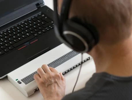 Person mit Kopfhörern sitzt vor Laptop; ein weißes Gerät mit vielen kleinen quadratischen Tasten liegt vor dem Laptop.