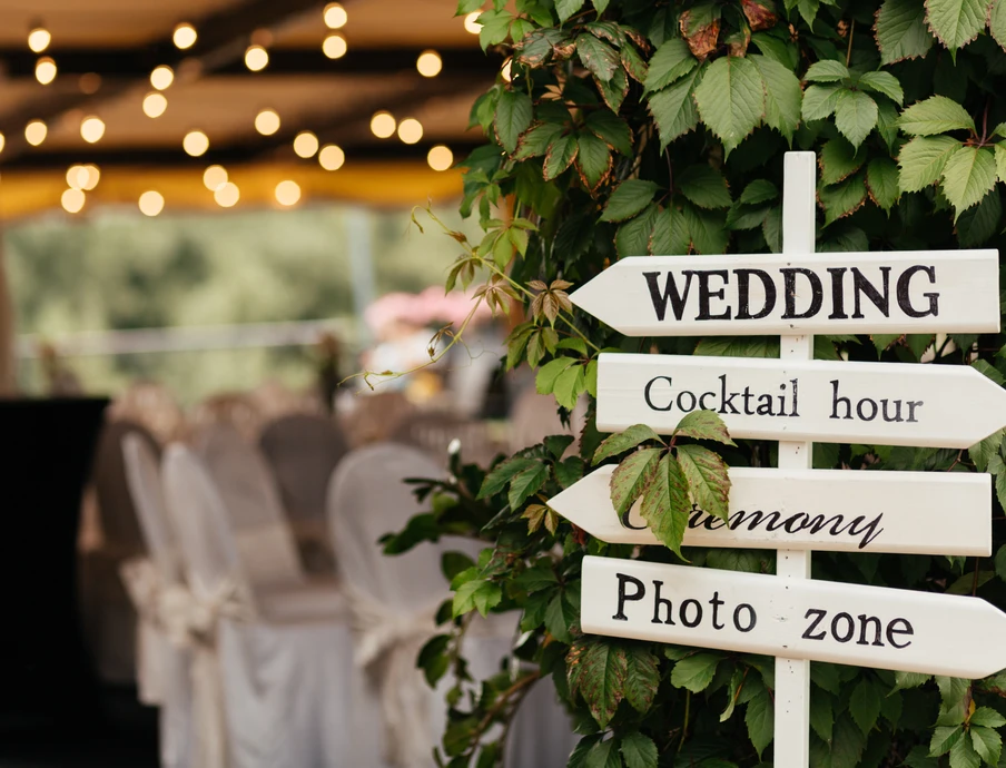Weißes Holzschild mit Pfeilen (WEDDING, Cocktail hour, Photo zone) an einer Grünpflanze; im Hintergrund Tische und Lichter.