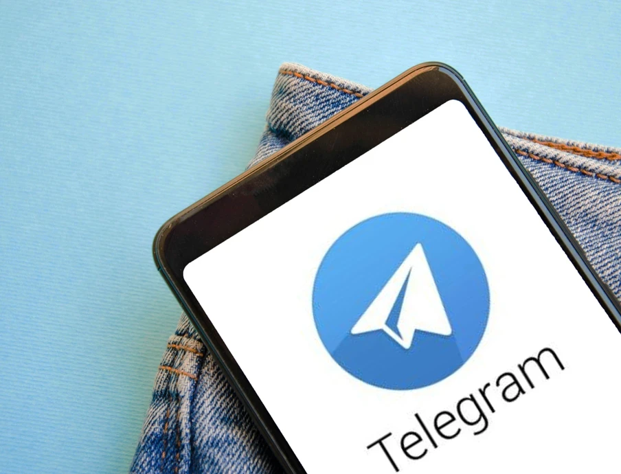 Smartphone mit Telegram-Logo (weißer Papierflieger auf blauem Kreis) auf blauem Hintergrund neben Jeansstoff.