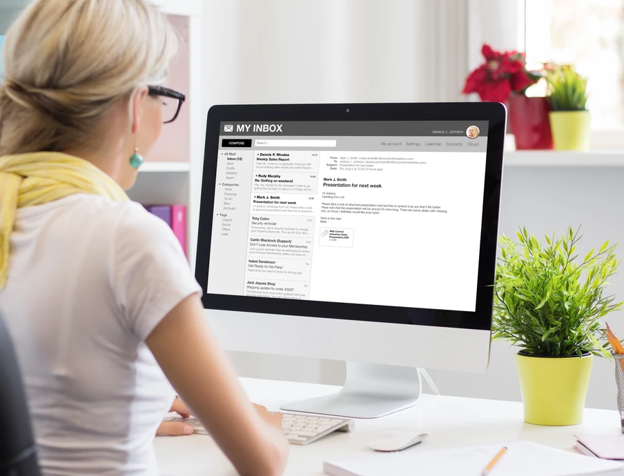 Frau mit Brille und gelbem Schal sitzt am Schreibtisch vor Computerbildschirm, der E-Mail-Posteingang sichtbar; Pflanze rechts.