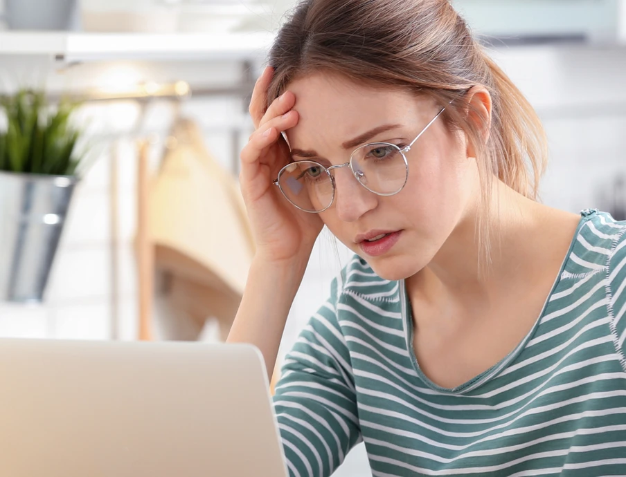 Frau mit Brille sitzt an einem Laptop, berührt Stirn mit der Hand; trägt gestreiftes Oberteil, helles Büro im Hintergrund.