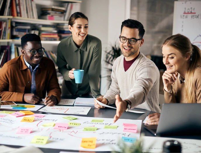 Vier Menschen sitzen um einen Tisch mit bunten Post-its, Notizbüchern, Laptop und einer Tasse.