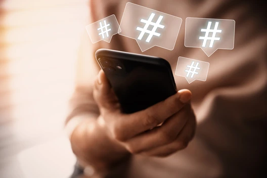 Person hält Smartphone, umgeben von schwebenden Hashtag-Symbolen in quadratischen Blasen.
