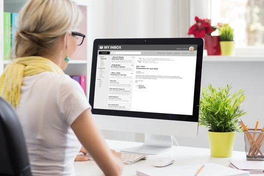 Frau mit Brille und gelbem Schal sitzt am Schreibtisch vor Computerbildschirm, der E-Mail-Posteingang sichtbar; Pflanze rechts.