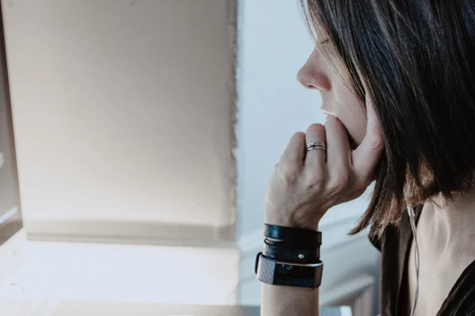 Person mit dunklem Haar sitzt vor zwei Monitoren mit Code; Hand am Kinn, Uhr am Arm.