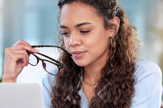 Frau mit lockigen Haaren hält eine Brille in der Hand und schaut auf einen Laptop.